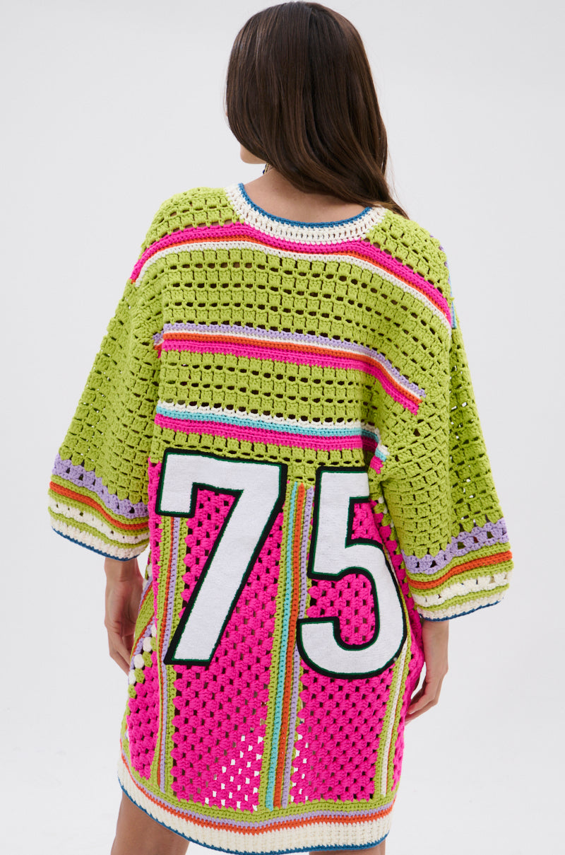 A person with long brown hair faces away, wearing the COURTSIDE OR SEASIDE CROCHET JERSEY DRESS—a loose crochet style with colorful stripes and a bold white and black number 75 on the back.