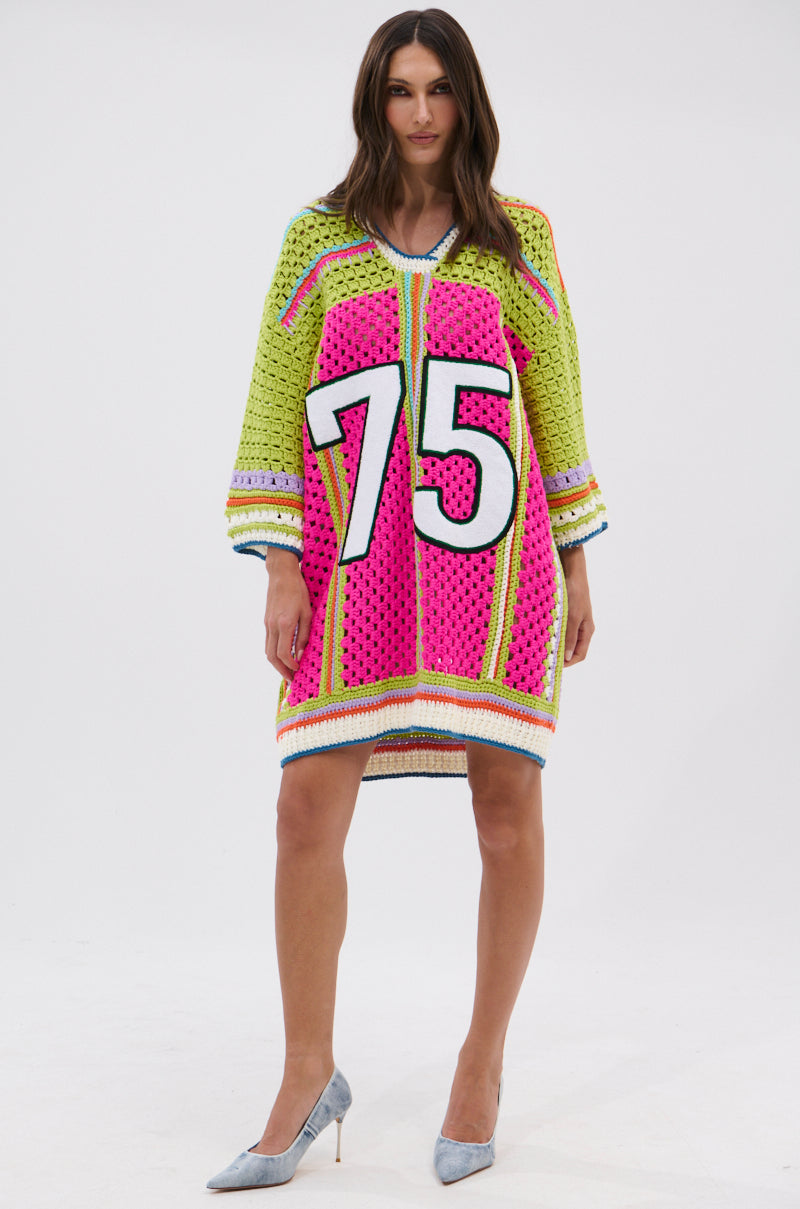 A woman faces forward in the COURTSIDE OR SEASIDE CROCHET JERSEY DRESS—an oversized, colorful sweater dress with bright pink, yellow, green, and blue patterns and a white 75 on the front. She completes the look with gray high heels.