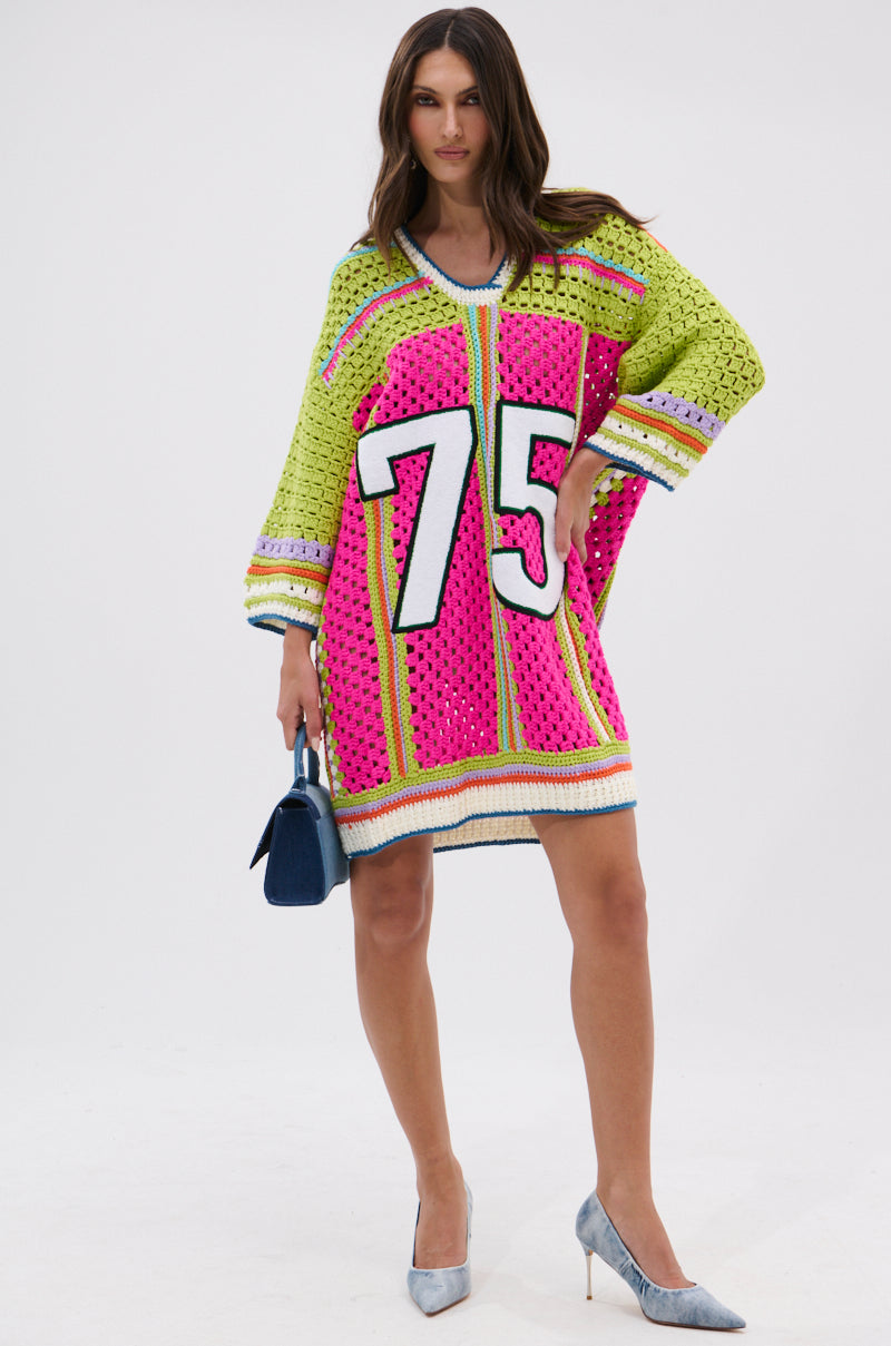 A woman models the COURTSIDE OR SEASIDE CROCHET JERSEY DRESS, a loose, colorful crochet piece featuring “75” on the front. She holds a small blue handbag and poses in silver heels against a plain white background.