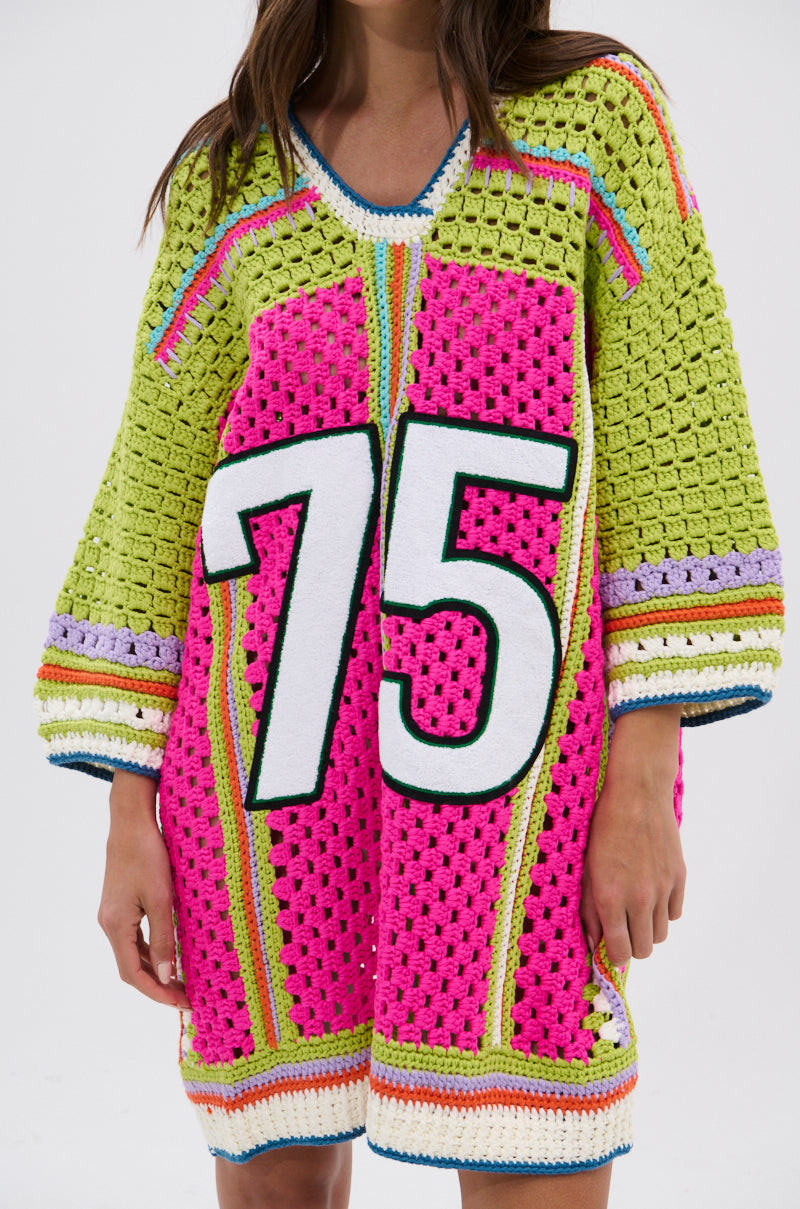 Someone wearing the COURTSIDE OR SEASIDE CROCHET JERSEY DRESS, an oversized, colorful sweater dress with wide sleeves, a bold white “75” outlined in black on the front, and vibrant pink, green, yellow, and multicolor accents.