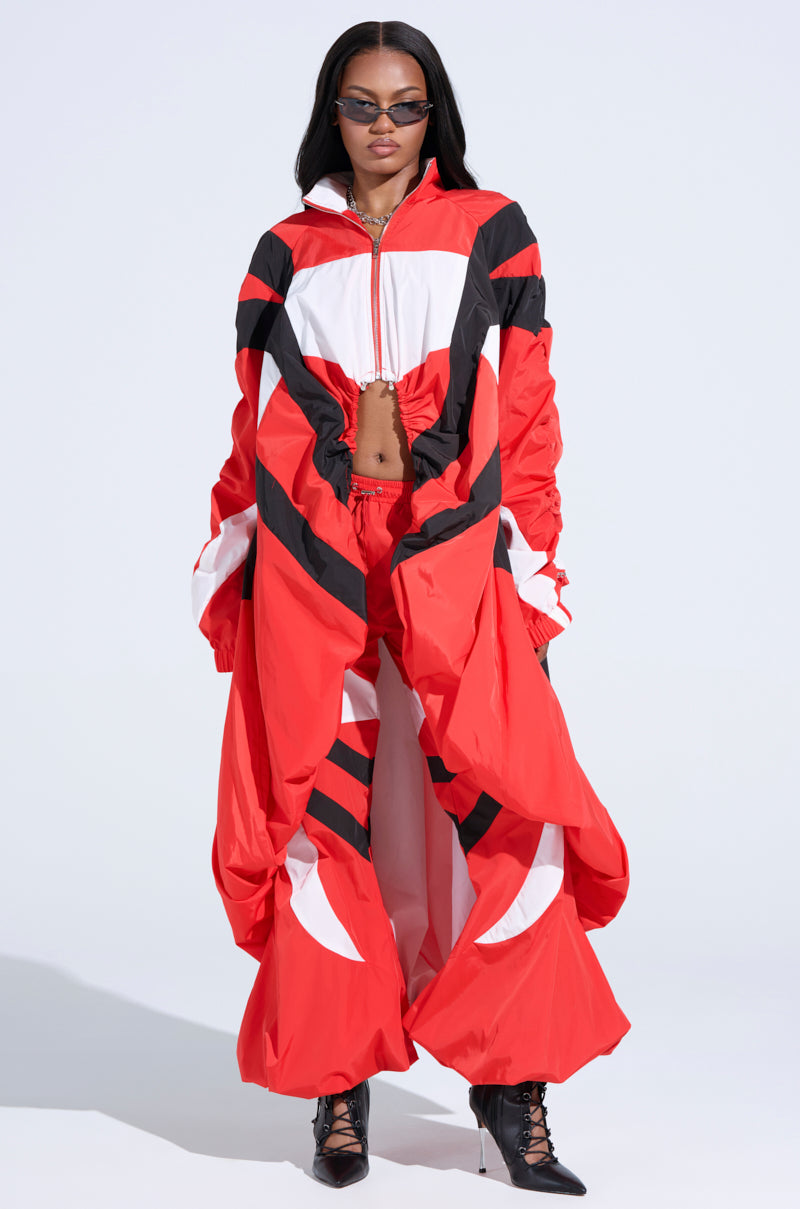 The individual wears an AKIRA Label ensemble, including the RED ALERT WINDBREAKER TRENCH in bold red and black with large white sections, featuring a dramatic drape and front zipper. The look is completed with sleek sunglasses and black pointed shoes.