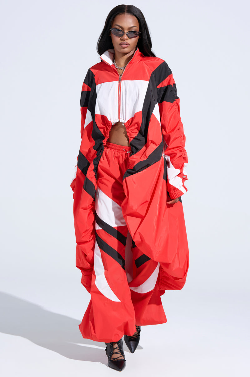 A person exudes confidence in a bold RED ALERT WINDBREAKER TRENCH featuring a red and black geometric pattern with a zipper front. Paired with sleek black sunglasses and high heels, they strut against a neutral background like they're in the editorial.