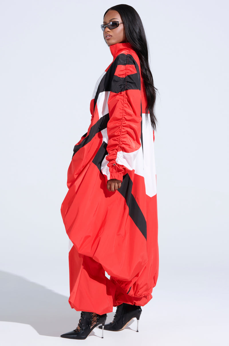 A person in large black sunglasses and a loose-fitting AKIRA Label geometric-patterned outfit strikes a sideways pose with one leg slightly forward. They have long black hair, wear pointed black ankle boots, and evoke the essence of the RED ALERT WINDBREAKER TRENCH.