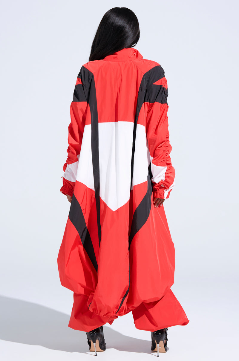 A person with long black hair, facing away, wears the bold RED ALERT WINDBREAKER TRENCH by AKIRA Label featuring black and white geometric patterns. The outfit includes matching pants and high-heeled shoes against a plain light background.