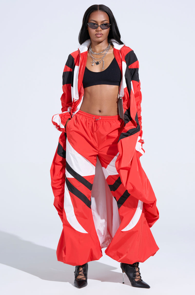 A woman confidently stands in a red and black athletic outfit, highlighting the RED ALERT WINDBREAKER JOGGERS with high-rise fit and color-blocked panels. She accessorizes with sunglasses, necklaces, and black ankle-strap heels against a plain white backdrop.