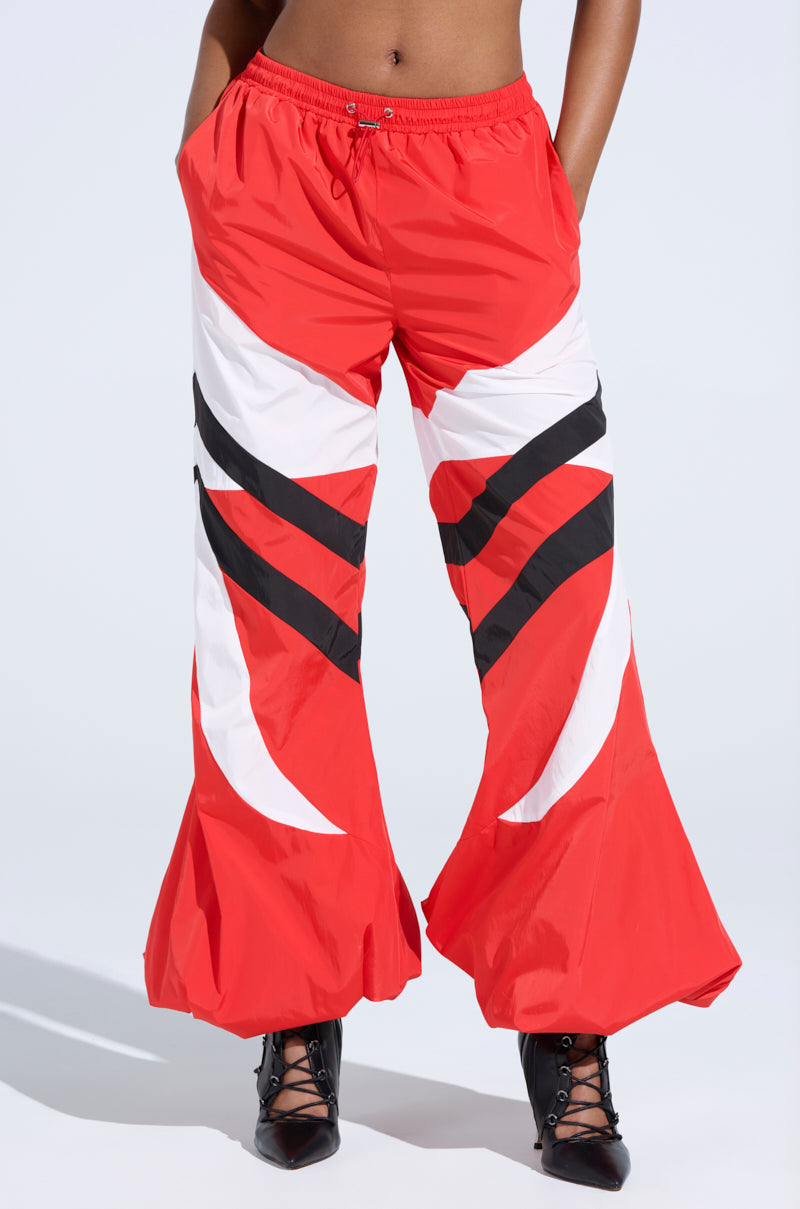 Someone is wearing the RED ALERT WINDBREAKER JOGGERS, featuring vibrant red, white, and black colors with bold diagonal stripes and flared wide legs. The high-rise fit is paired with black pointy lace-up shoes against a plain light backdrop.