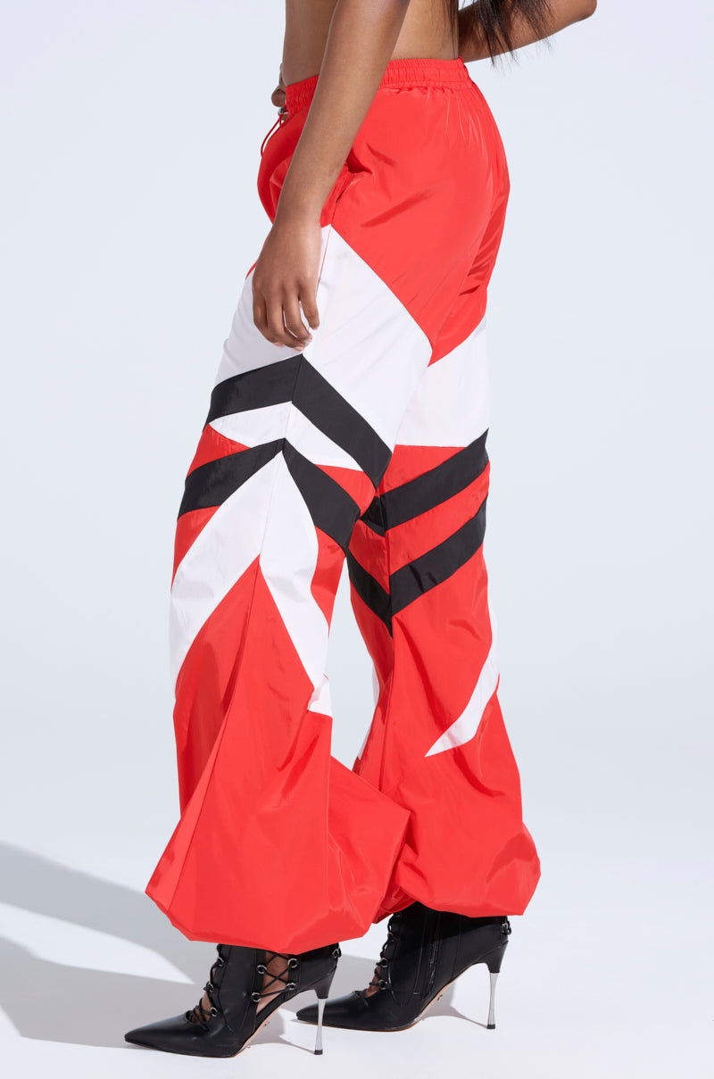 Someone is wearing wide-legged pants in striking red, white, and black geometric patterns reminiscent of RED ALERT WINDBREAKER JOGGERS. Paired with sleek high-heeled black boots, the high-rise design enhances elegance as they pose against a plain background.