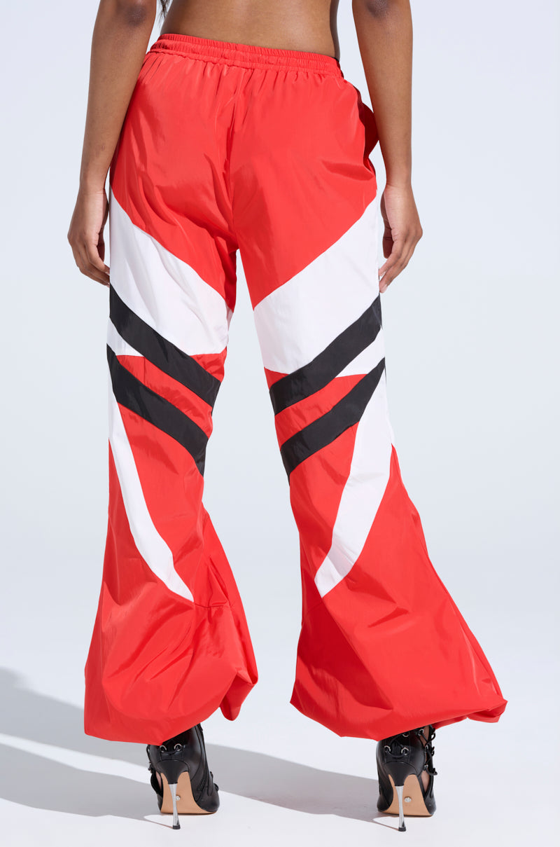 The image displays someone wearing Red Alert Windbreaker Joggers adorned with striking white and black chevron patterns on a vivid red, relaxed fit. They're stylishly paired with black high heels on a plain backdrop, highlighting the bold outfit.