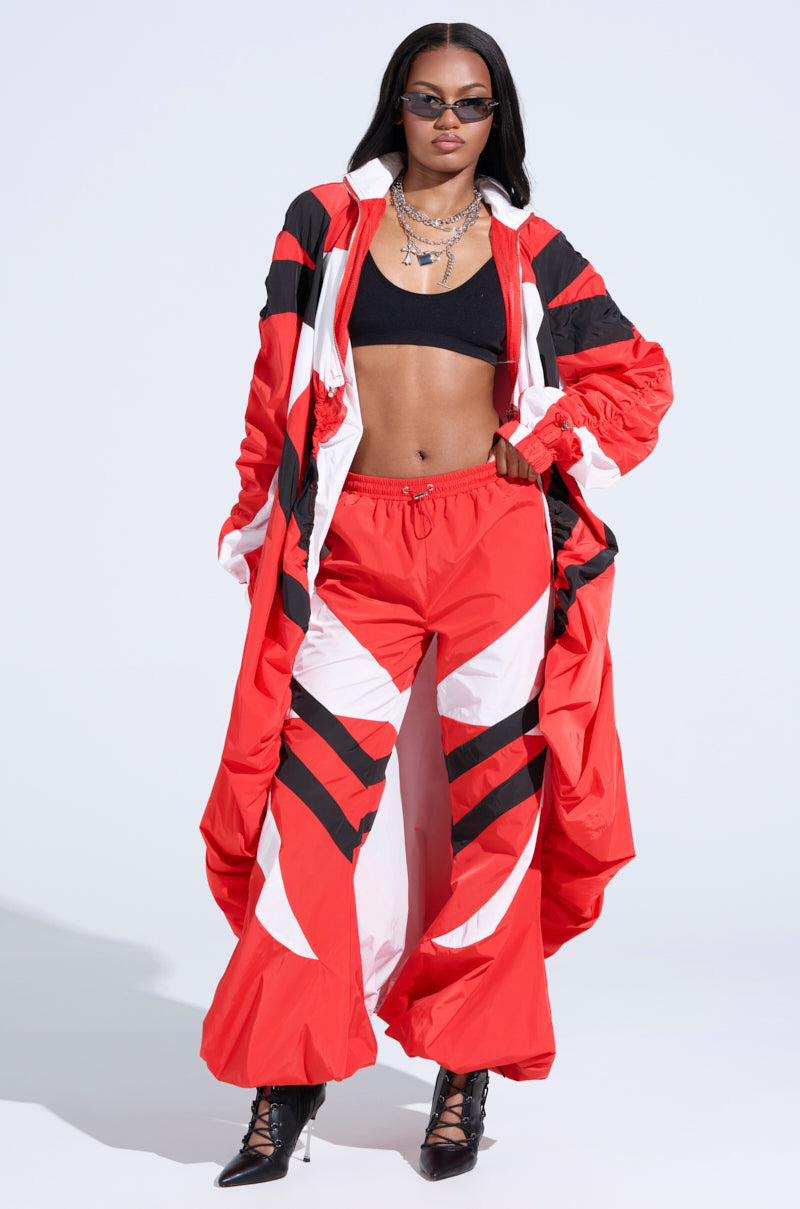 Against a plain background, a person confidently poses in a bold red and black geometric-patterned long coat with color-blocked panels, paired with high-rise RED ALERT WINDBREAKER JOGGERS, a black crop top, and heeled boots. Sunglasses and layered necklaces complete the striking look.