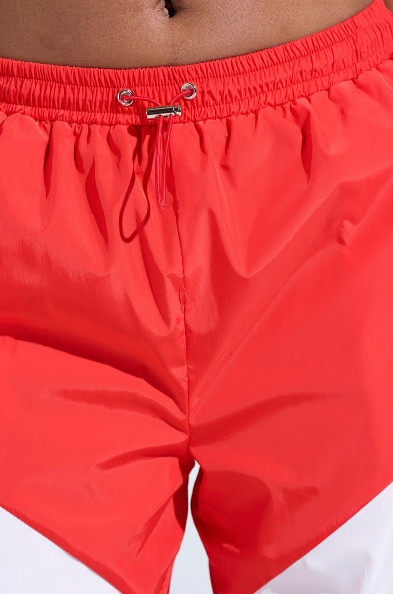Close-up of a person in RED ALERT WINDBREAKER JOGGERS with an elastic waistband and metal eyelets. The shiny, bright red fabric features color-blocked panels, offering a stylish flair with a slightly relaxed fit. The focus highlights the midsection's vibrant design.