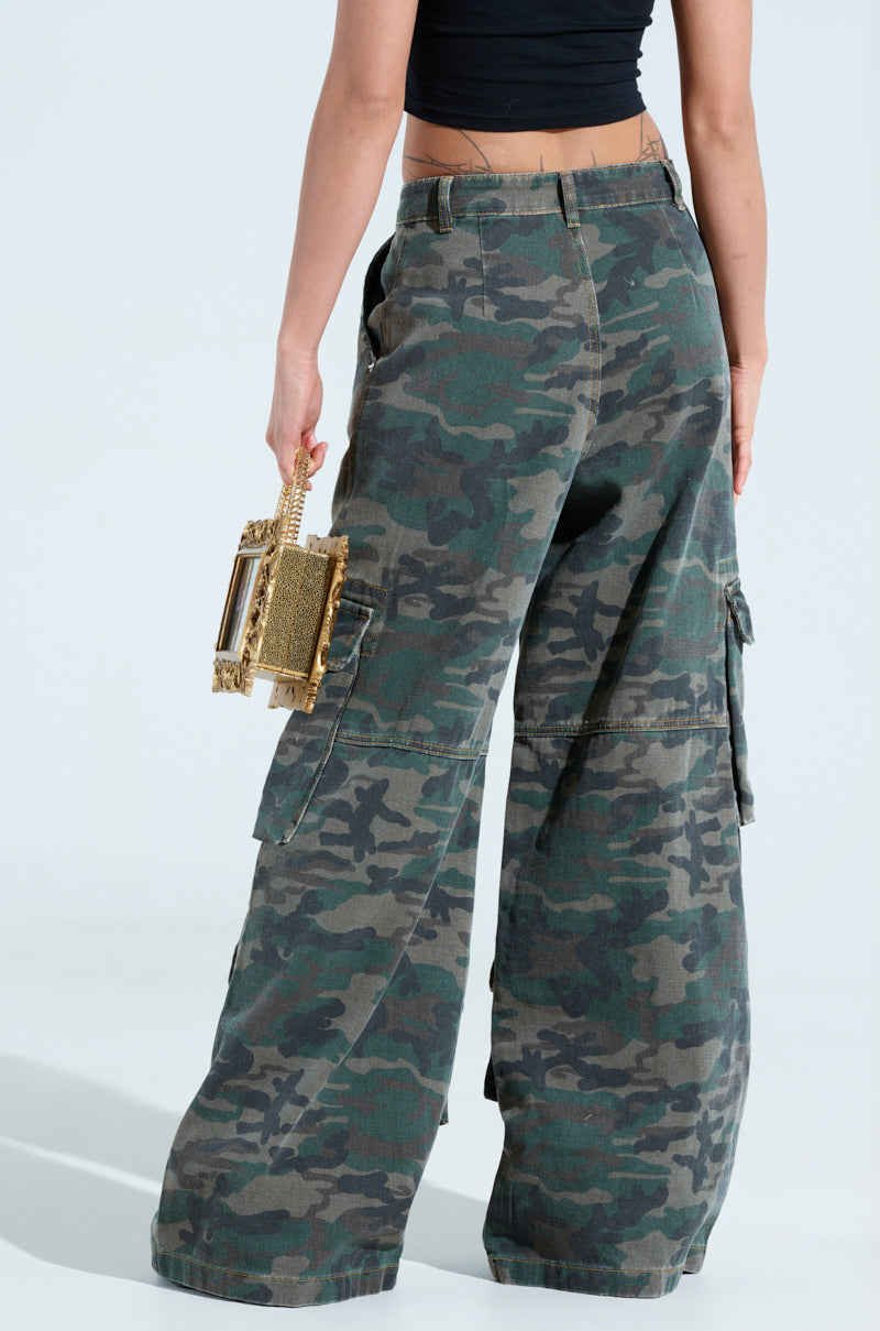 A person in a black crop top and NOT SO BASIC CAMO CARGO PANTS with wide-leg style holds a small gold clutch against a light, neutral background.