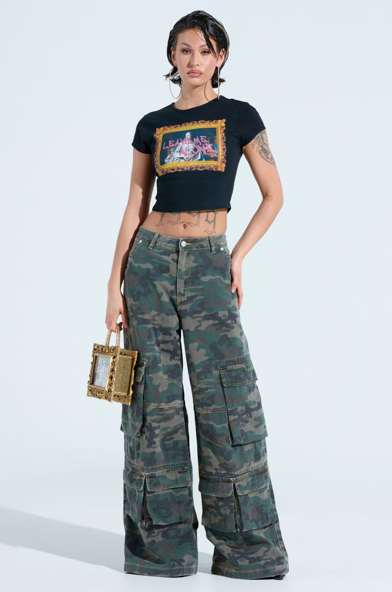With a slicked-back hairstyle, the individual confidently wears a black crop top with graphic design and NOT SO BASIC CAMO CARGO PANTS featuring a wide-leg silhouette. Tattoos adorn their arms and torso as they hold a small rectangular handbag against the plain background.