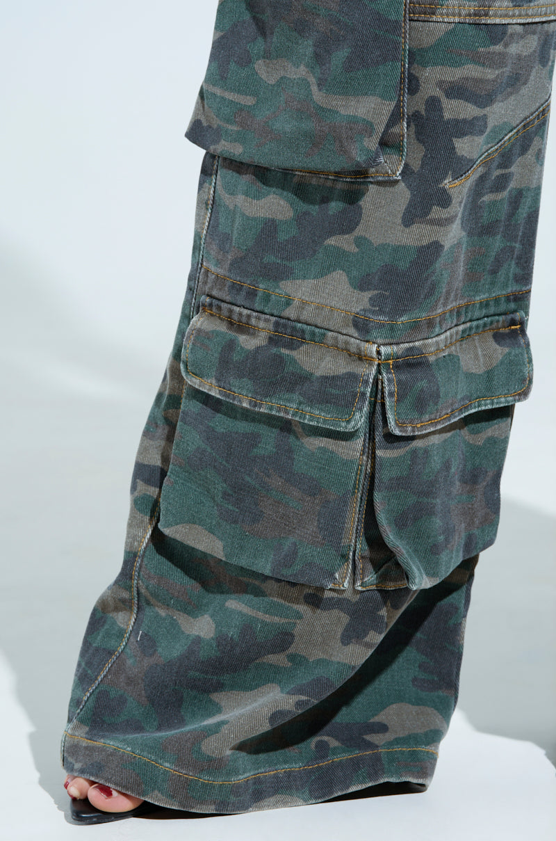 A close-up shows someone in NOT SO BASIC CAMO CARGO PANTS by AKIRA Label with wide legs and large pockets. The slightly oversized fit covers most of the foot, revealing just the shoe tip.