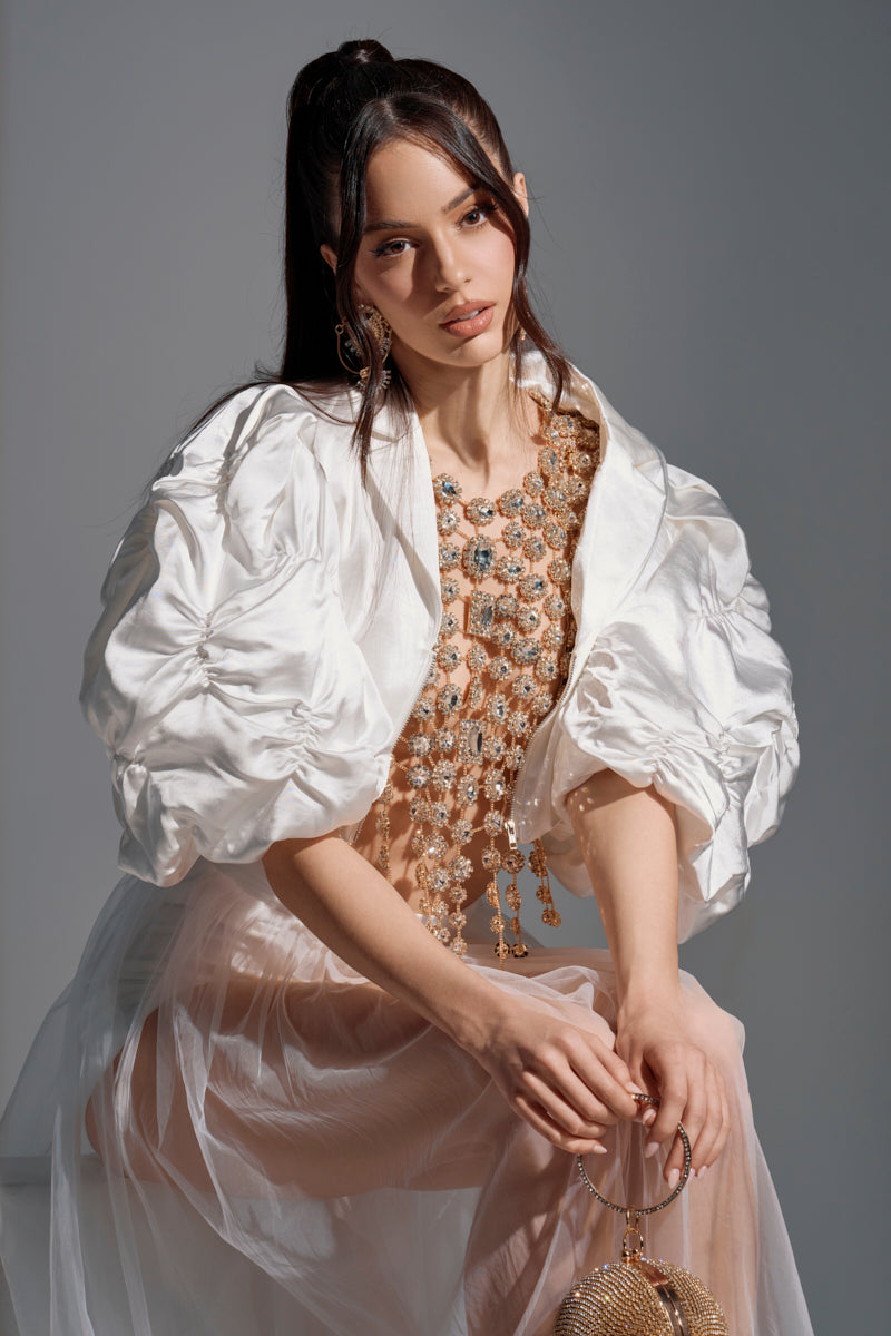 A woman with long dark hair in a high ponytail wears the HERE FOR A GOOD TIME RUFFLE SLEEVE BLAZER, styled with a sheer embellished top and light skirt. She sits gracefully, holding a sparkling round handbag in one hand.