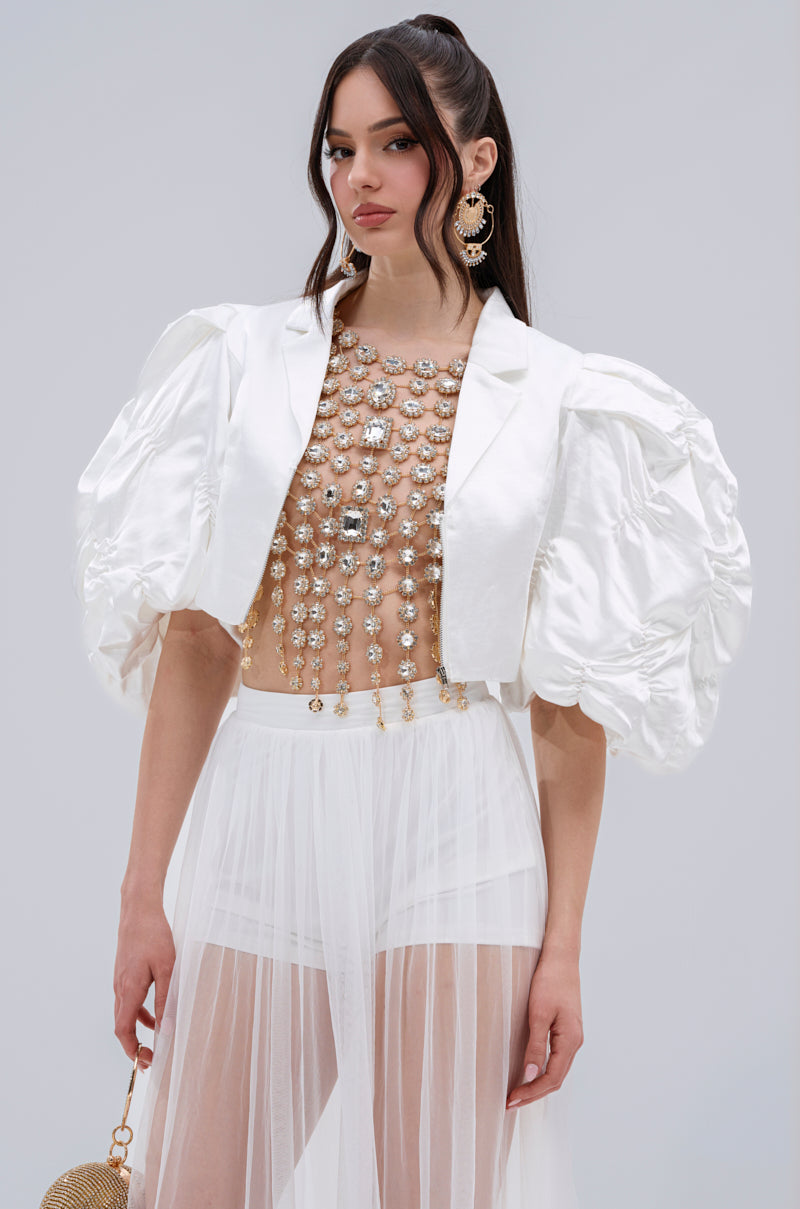 A woman in the HERE FOR A GOOD TIME RUFFLE SLEEVE BLAZER with puff sleeves wears it over a sheer, pearl and jewel-embellished dress, standing against a plain light background and holding a small round gold bag.