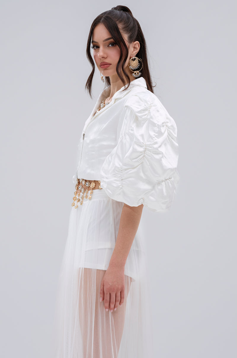 A woman with long dark hair in a high ponytail models the HERE FOR A GOOD TIME RUFFLE SLEEVE BLAZER with voluminous puff sleeves, a sheer white skirt, statement earrings, and a gold belt against a plain light grey background.
