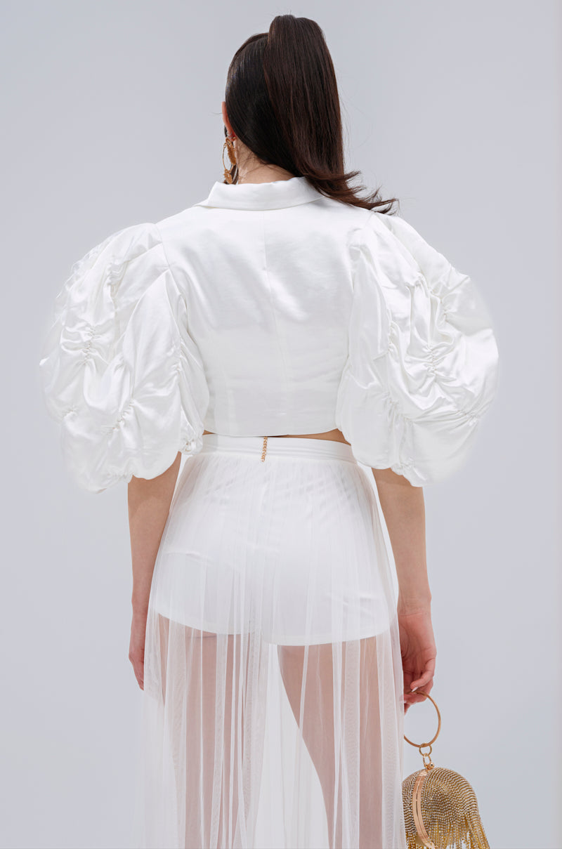 A woman faces away, wearing the HERE FOR A GOOD TIME RUFFLE SLEEVE BLAZER—a white satin piece with dramatic puffy sleeves—styled with a sheer skirt over shorts, a gold mesh handbag, and her dark hair in a high ponytail.
