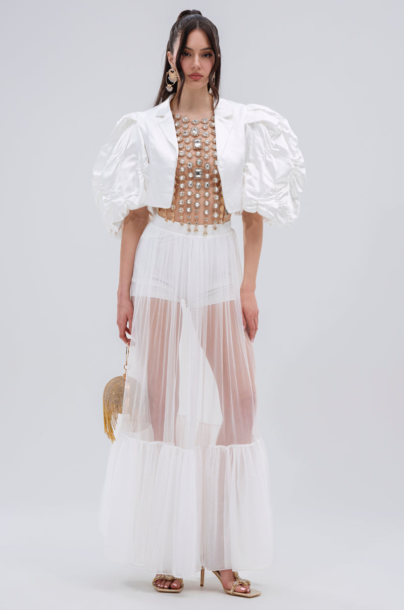 A woman wears a sheer white skirt, high-waisted white briefs, a pearl-embellished nude top, and the HERE FOR A GOOD TIME RUFFLE SLEEVE BLAZER. She accessorizes with a gold fringed purse and gold heels.