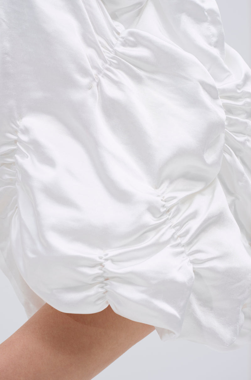 Close-up of white, ruched satin fabric with gathered seams and a soft texture; part of the HERE FOR A GOOD TIME RUFFLE SLEEVE BLAZER worn by a person, with some skin visible at the bottom edge.