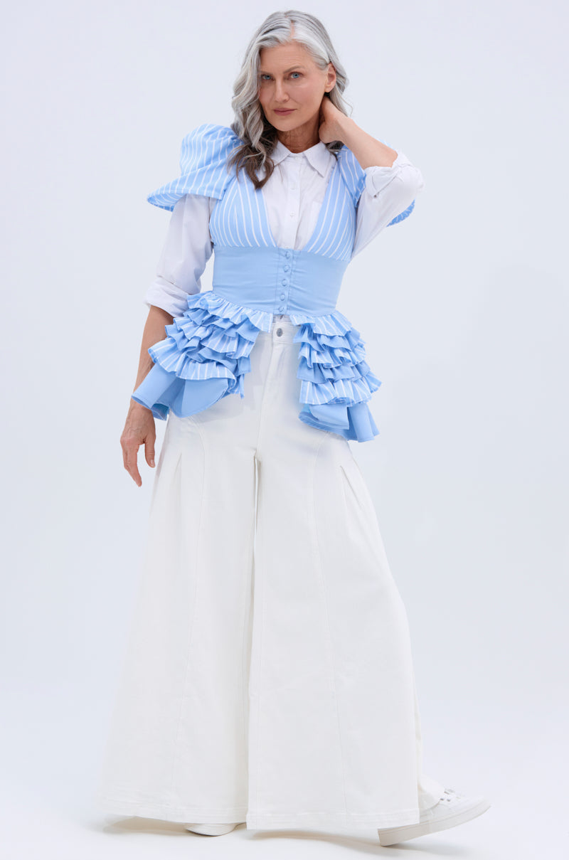 A woman with long gray hair poses in TRENDY AND COOL WHITE WIDE LEG DENIM PANT, styled with a white button-up shirt and a light blue and white striped vest with ruffled waist accents. The background is plain and light-colored.