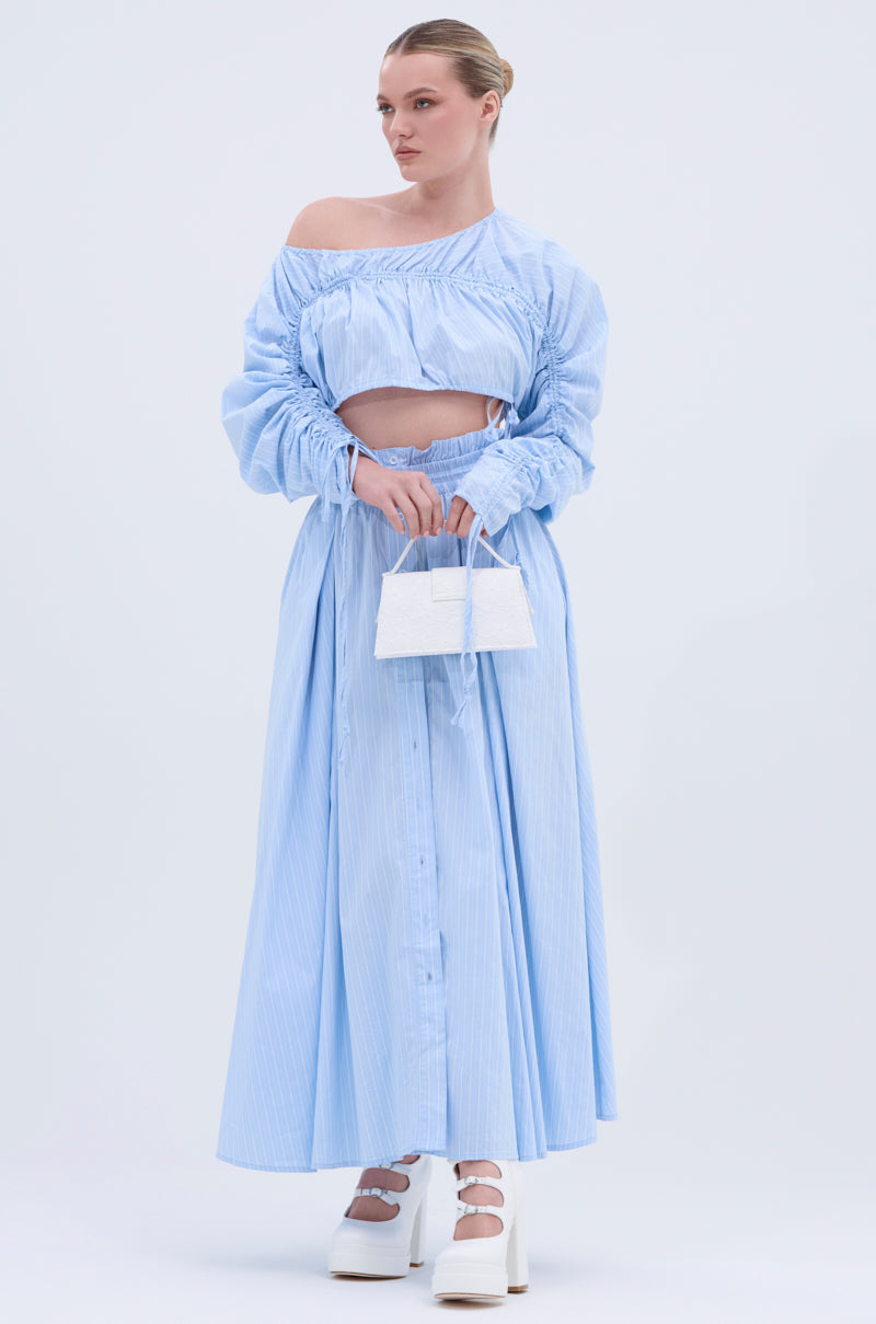 A woman wears the TALK TO ME MAXI SKIRT, a light blue, off-the-shoulder, long-sleeve crop top and matching A-line maxi skirt. She accessorizes with a white handbag and platform shoes, her hair neatly tied back.