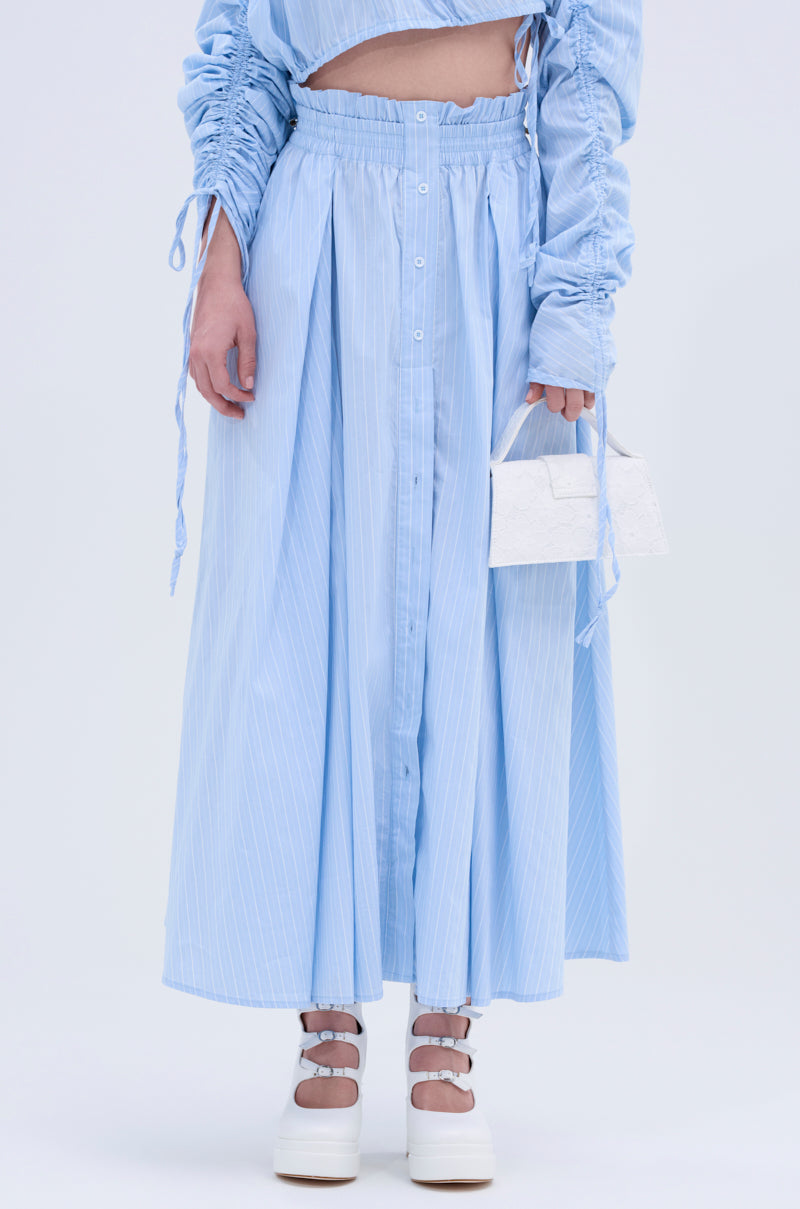 Someone is wearing the TALK TO ME MAXI SKIRT in light blue pinstripes with an A-line silhouette, a matching long-sleeve top, white platform shoes, and holding a small white handbag. Their face is not visible.