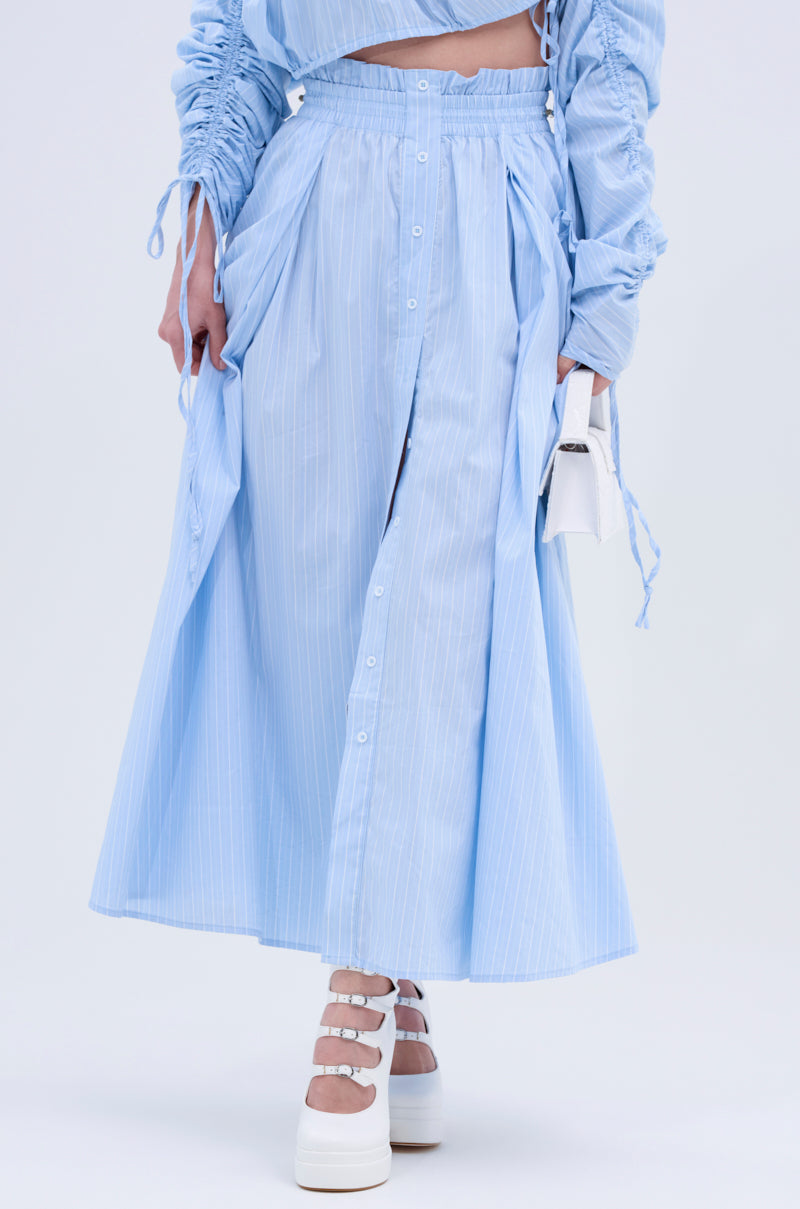 Someone wears the TALK TO ME MAXI SKIRT, a light blue pinstripe ankle-length skirt with ruched details, paired with a matching ruched top, a small white handbag, and chunky white platform heels.