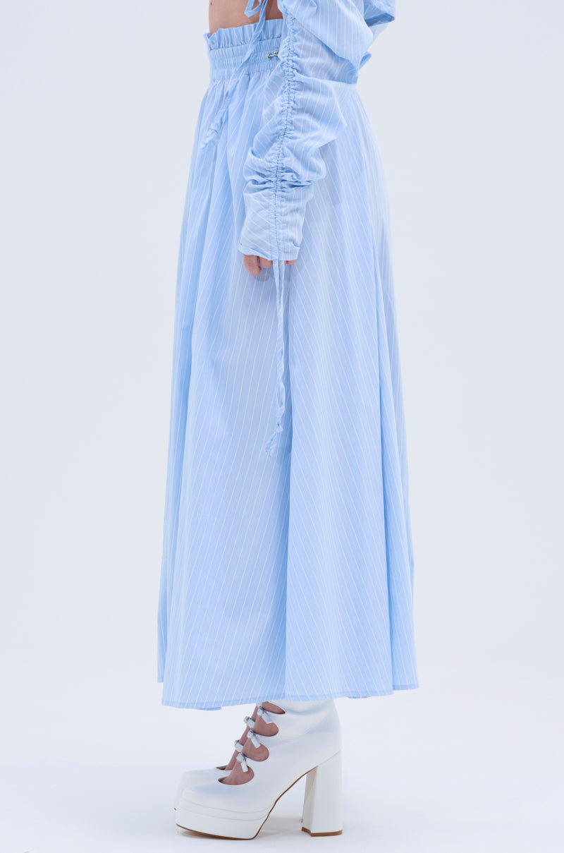 A model in the TALK TO ME MAXI SKIRT, light blue with pinstripes and an A-line silhouette, matched with a ruched-sleeve top and white chunky cut-out heels, stands against a plain white background.