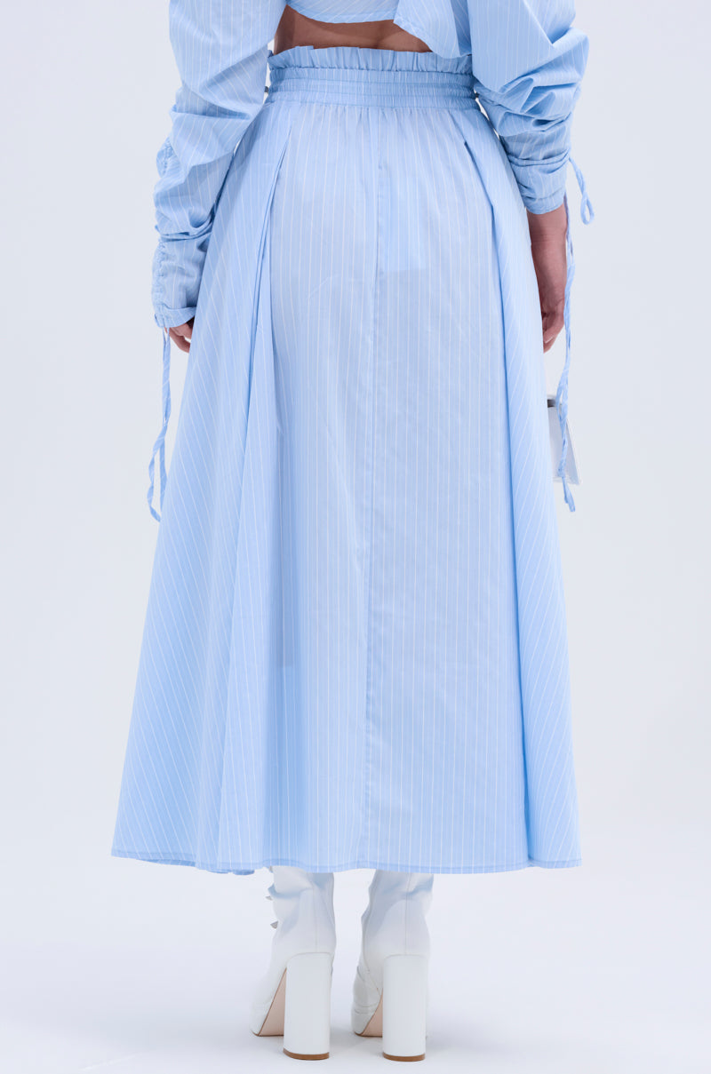 A person models the TALK TO ME MAXI SKIRT—light blue, ankle-length with subtle pinstripes, slight pleats, gathered waistband, and A-line silhouette—paired with white heeled boots, shown from the back against a plain background.