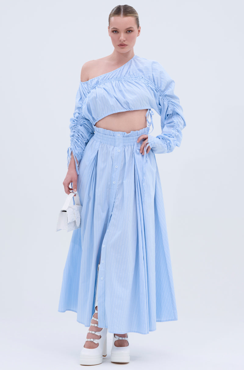 A woman wears the TALK TO ME MAXI SKIRT, a light blue, pinstripe A-line maxi skirt with an asymmetrical crop top. She accessorizes with a small white handbag and white platform shoes against a light background.