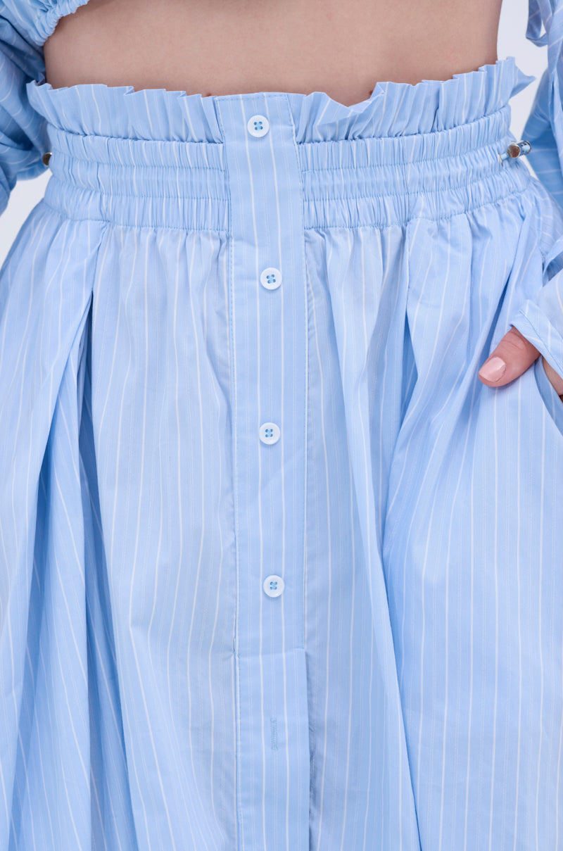 Close-up of the TALK TO ME MAXI SKIRT: a light blue pinstriped maxi with an elastic ruched high waistband, white front buttons, and side pockets. A hand rests in the pocket, highlighting its A-line silhouette.