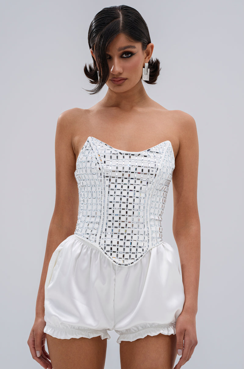 A woman in the WHITE HOT EMBELLISHED CORSET, with a structured grid-patterned mirrored bodice and gathered satin shorts, stands against a light gray background, wearing wavy dark pigtails and square earrings.