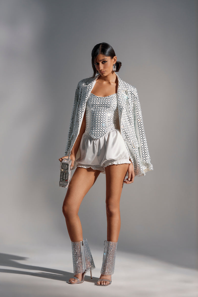 A woman stands in a studio, wearing the WHITE HOT EMBELLISHED CORSET with mirrored details, paired with white satin shorts and clear heels. Holding a retro phone, she gazes confidently at the camera.