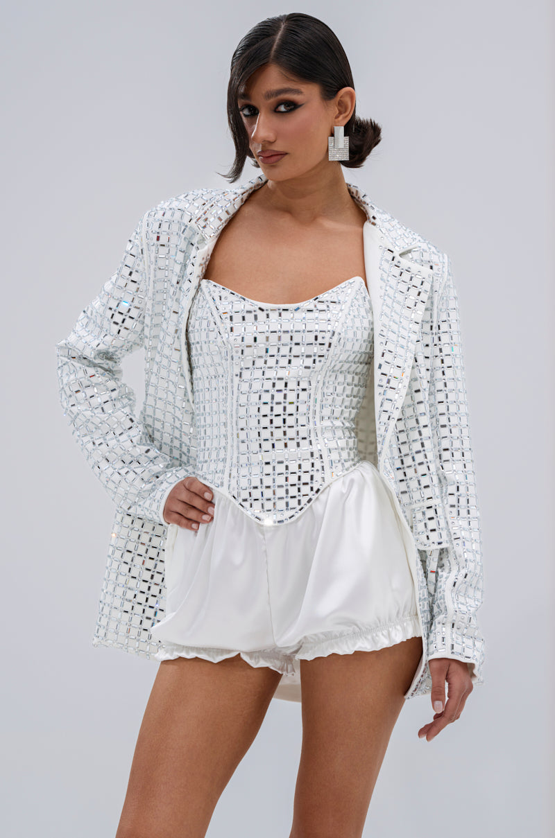 A woman exudes confidence in the WHITE HOT EMBELLISHED BLAZER, featuring a notch lapel collar and mirrored details, styled over a matching corset and voluminous satin shorts with geometric earrings, sleek side-parted hair, and bold makeup.
