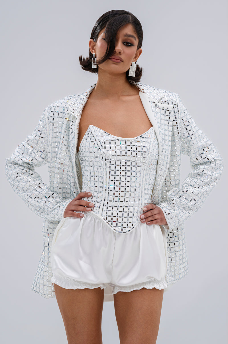 A woman poses confidently in a stylish, futuristic outfit featuring the WHITE HOT EMBELLISHED BLAZER with mirrored details and a notch lapel, paired with a matching corset and bubble shorts against a light gray background.