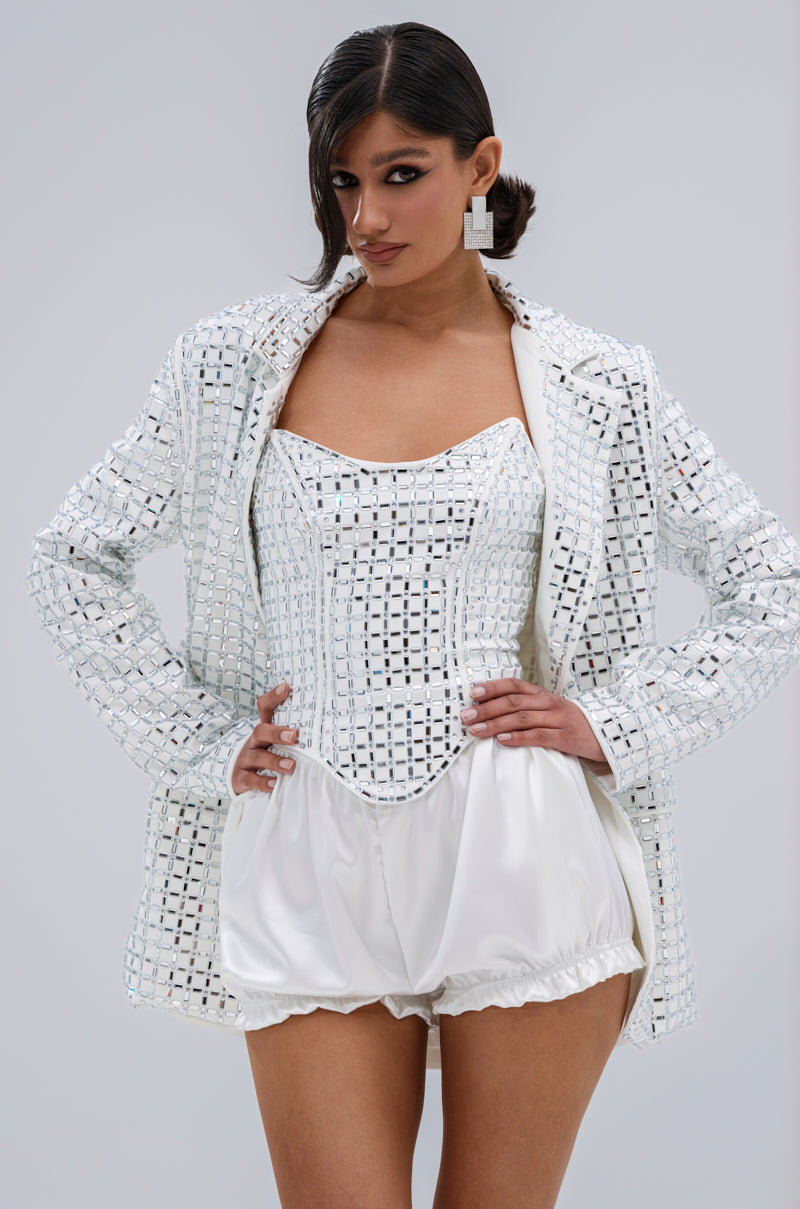 A woman poses confidently in the WHITE HOT EMBELLISHED BLAZER, featuring mirrored embellishments and a notch lapel collar, styled over a matching corset and satin balloon shorts with bold earrings and side-swept hair.