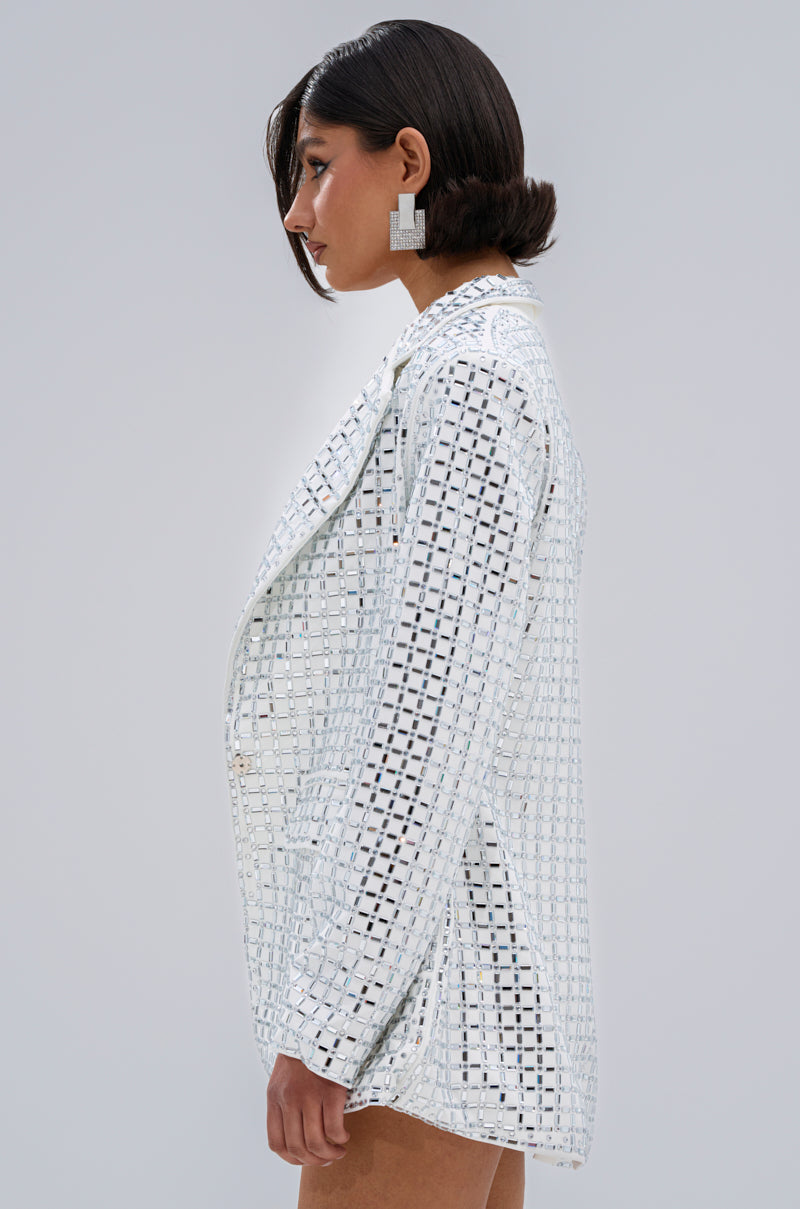A woman with short dark hair models the WHITE HOT EMBELLISHED BLAZER, featuring a notch lapel collar and mirrored details, along with geometric silver earrings, posing in profile against a plain light gray background.
