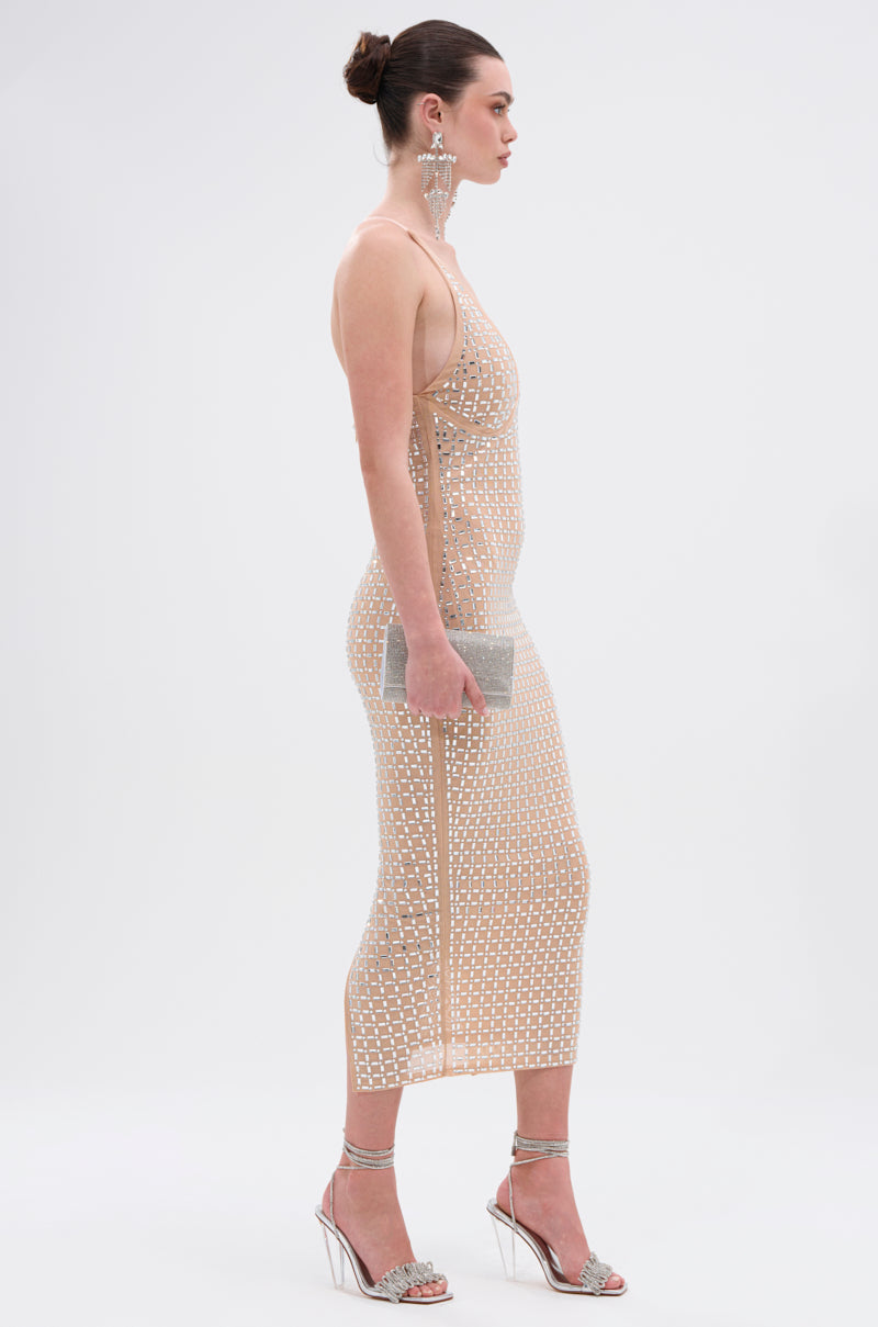 A woman in the QUEEN OF EVERYTHING MIDI DRESS—a nude, fitted, sleeveless dress with a white grid pattern and sheer double-lined mesh—stands in profile holding a silver clutch, wearing strappy silver heels, and her hair is in a neat bun. 3