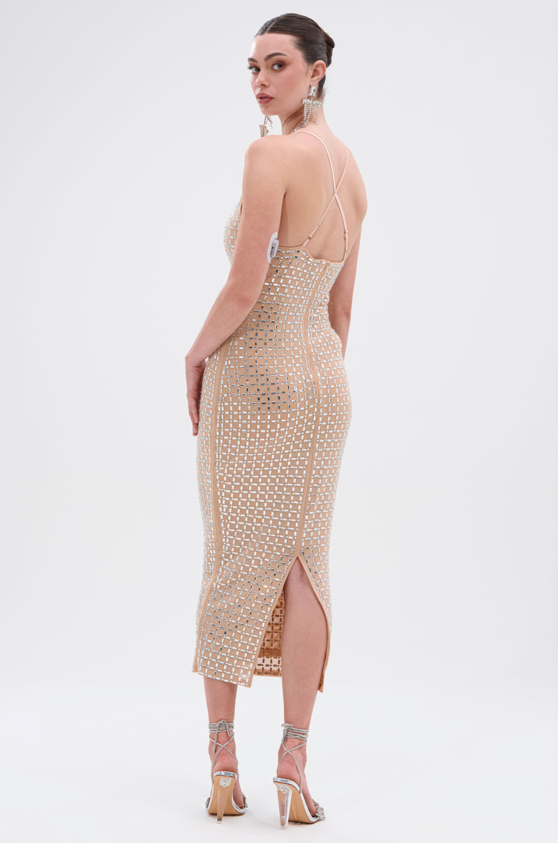 A woman stands facing backward in the QUEEN OF EVERYTHING MIDI DRESS, a beige fitted midi with mirrored embellishments, thin straps, and a back slit. She wears silver heels and statement earrings, her hair styled back. 4