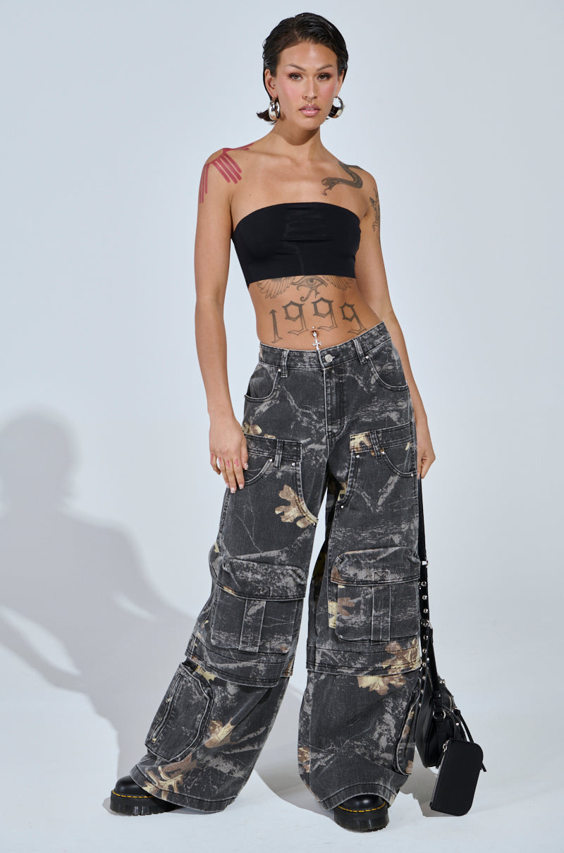A woman with short hair and tattoos wears a black bandeau top and the DOWN AND DIRTY CARGO CAMO PANT, paired with a black handbag. She stands confidently against a light gray background.