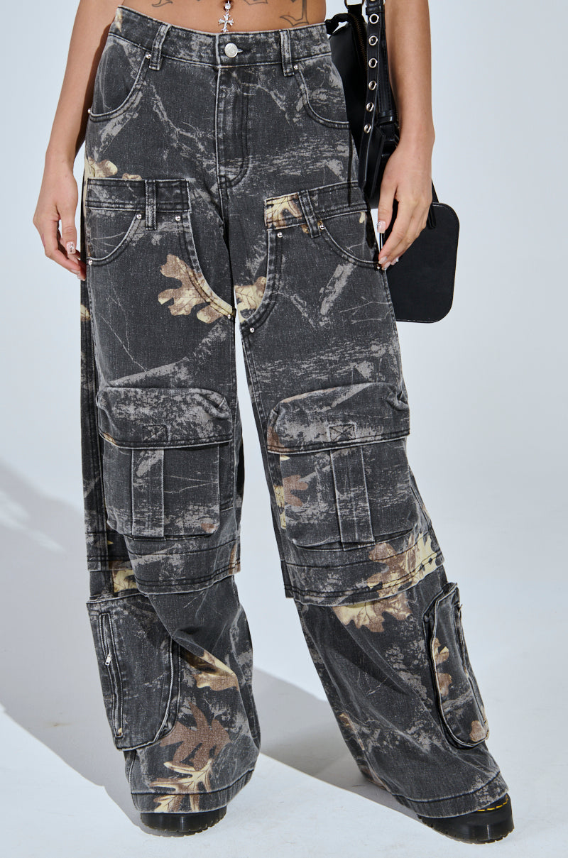 Wearing the DOWN AND DIRTY CARGO CAMO PANT, featuring a high-rise fit, black camo print, wide leg silhouette, and multiple large pockets, a person stands with one hand visible holding a black bag.