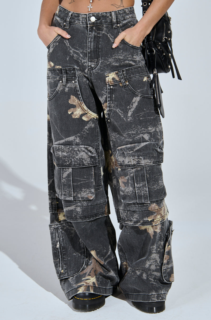 Wearing the DOWN AND DIRTY CARGO CAMO PANT in loose black camo with a faded abstract pattern, large pockets, and ankle zippers, the person keeps their hands in their pockets and carries a black bag on one arm.