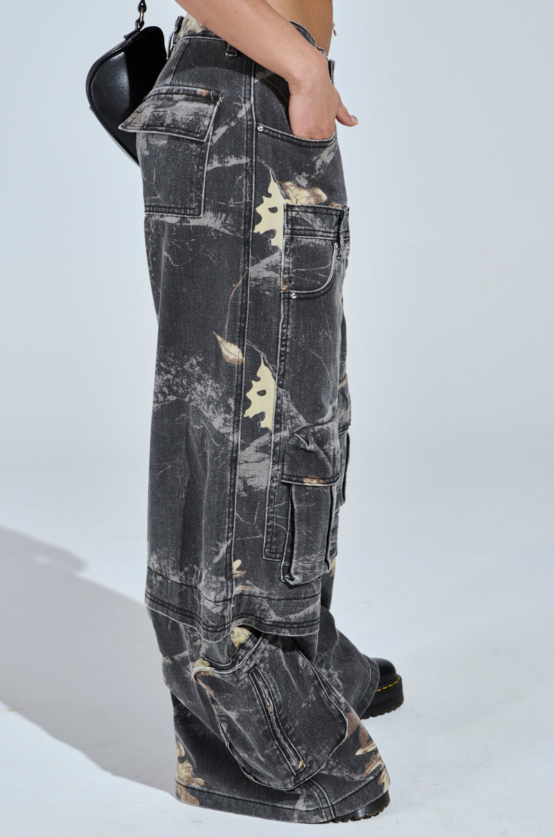 A person stands sideways wearing the DOWN AND DIRTY CARGO CAMO PANT in loose-fitting black and gray camo with a wide leg silhouette, faded distressed look, and multiple pockets, paired with a small black bag.