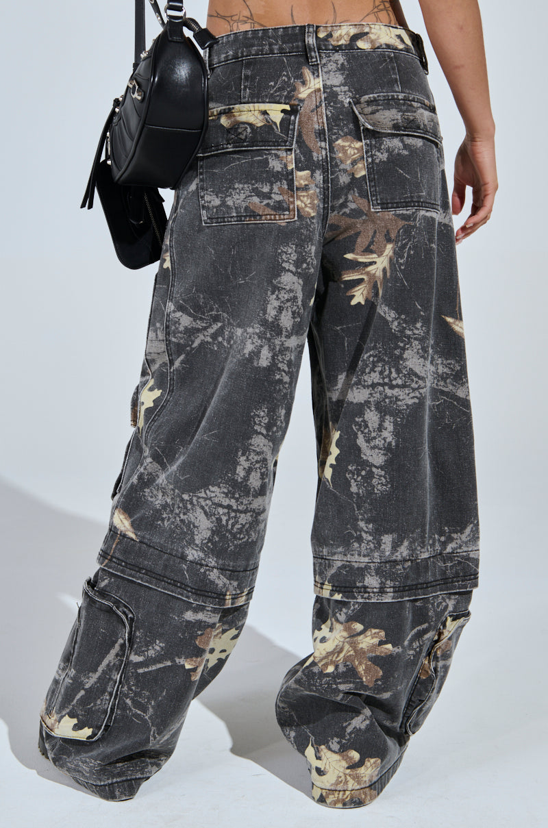 Wearing the DOWN AND DIRTY CARGO CAMO PANT in high-rise black camo with abstract beige and brown leaf print, this person stands facing away, carrying a black shoulder bag that highlights the wide leg silhouette.