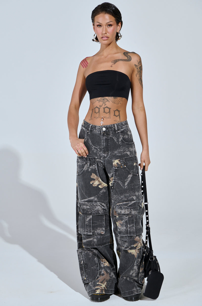 A woman with tattoos poses against a plain background, wearing a black bandeau top and the DOWN AND DIRTY CARGO CAMO PANT with a wide leg silhouette. She holds a small black bag and has short, slicked-back hair.