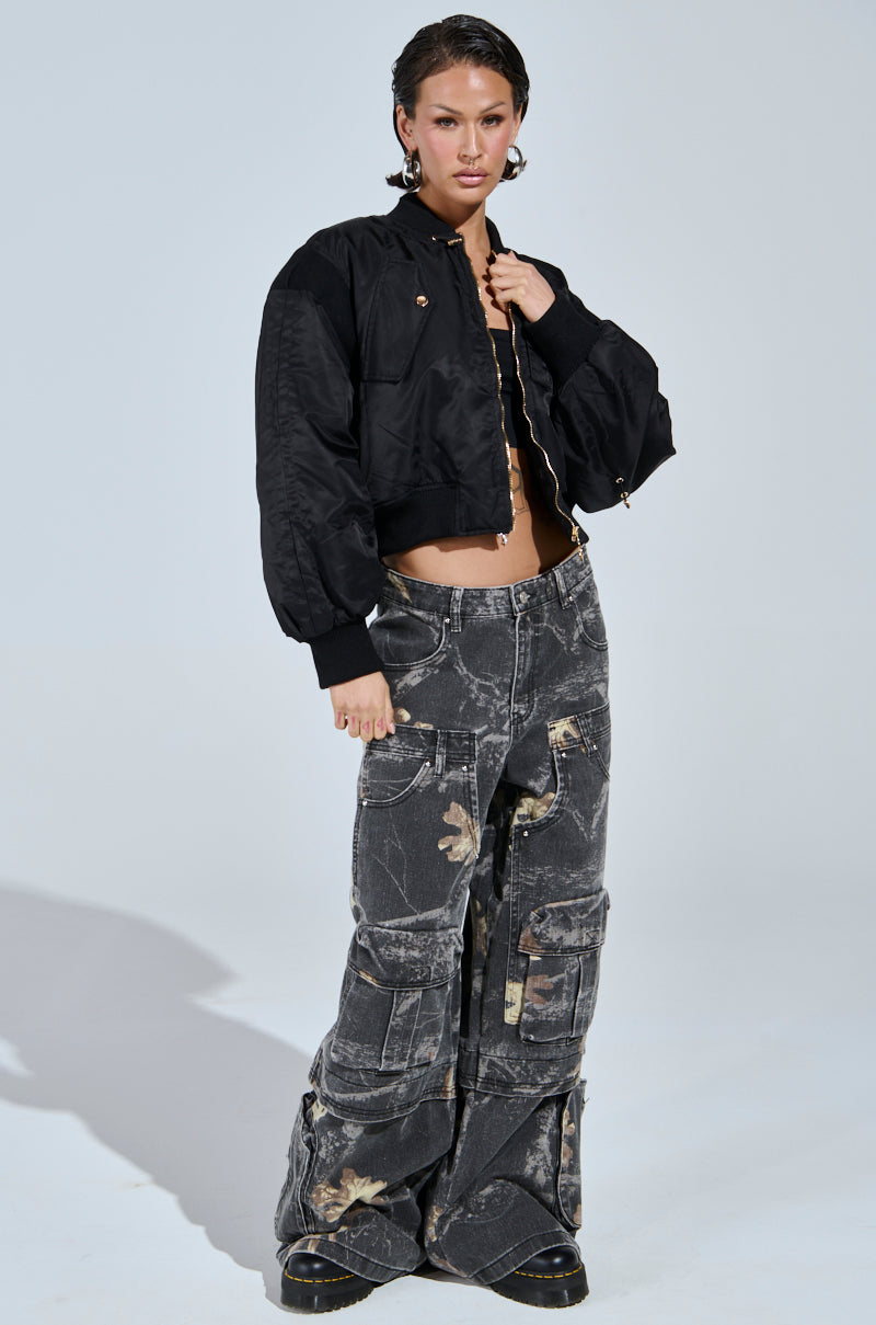 A person stands confidently in a cropped black bomber jacket and the DOWN AND DIRTY CARGO CAMO PANT, featuring a high-rise wide leg camo silhouette with a faded pattern. They finish the look with large hoop earrings and short, wet-look hair.