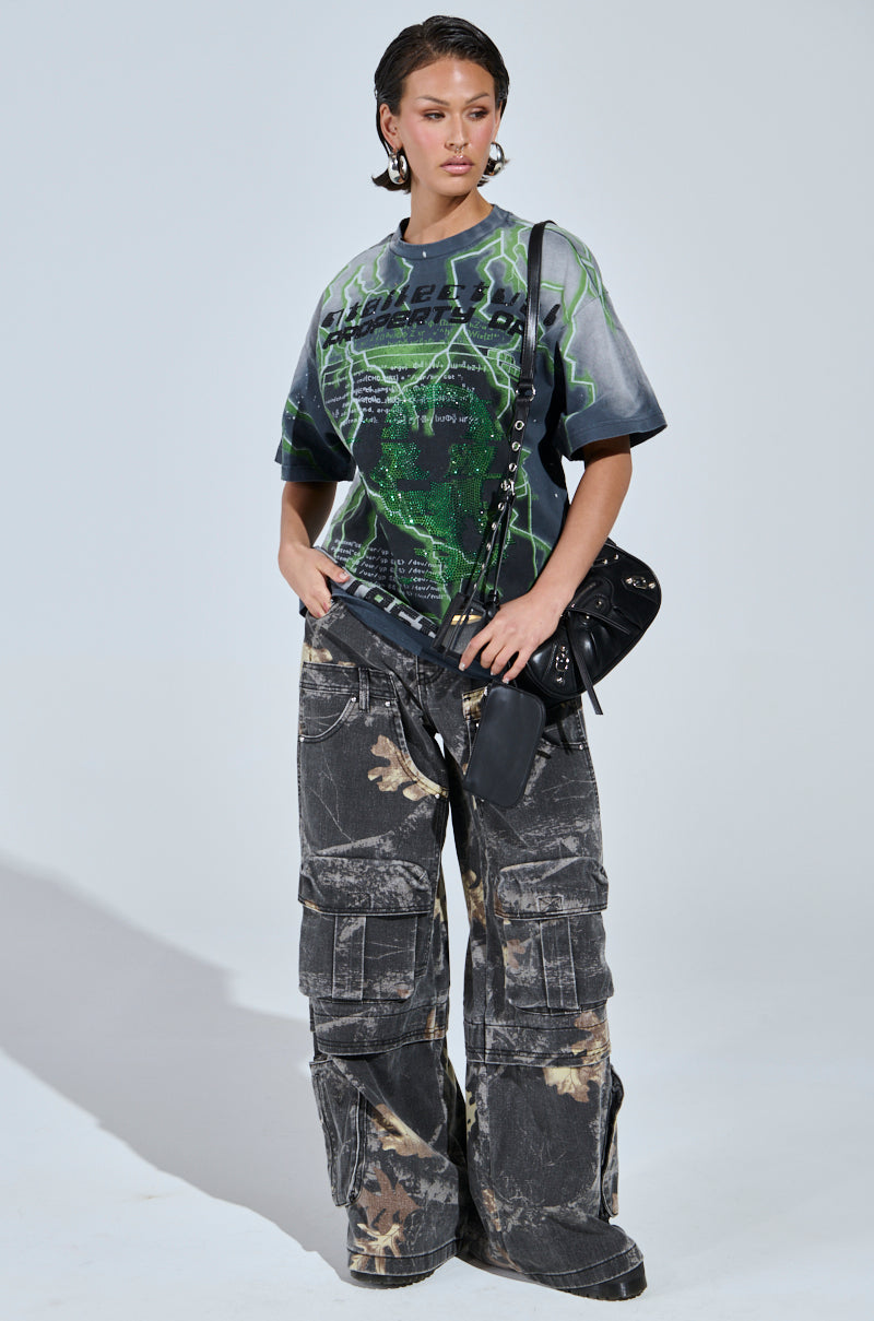 A woman stands against a plain background wearing a graphic tee, holding a black shoulder bag, and sporting the DOWN AND DIRTY CARGO CAMO PANT with a high-rise wide leg silhouette and distressed acid-wash finish.