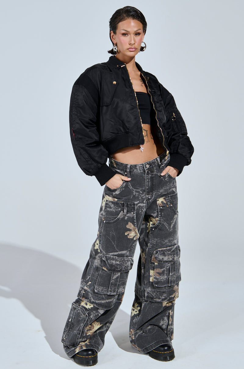 A person stands confidently in a black cropped jacket, black tube top, and the DOWN AND DIRTY CARGO CAMO PANT featuring a wide leg and faded patchwork pattern. They wear black shoes, have short hair, and accessorize with hoop earrings.