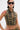 A person with short dark hair, visible arm and torso tattoos, a nose ring, and dark sunglasses wears the DARIA STUDDED CAMO SIDE LACE TOP featuring leopard print, gold studs, and lace-up sides.