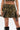A person wears the DARIA STUDDED CAMO MINI SKIRT with gold-tone studs, a matching camo crop top, and black boots, showing off a thigh tattoo and a belly button piercing.